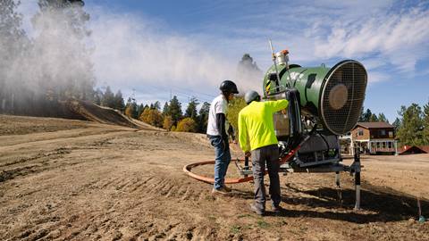 Two adults, one in a white shirt and the other in a bright yellow, inspecting a fan gun snow machine on-hill during a sunny fall day for the snow making test day.