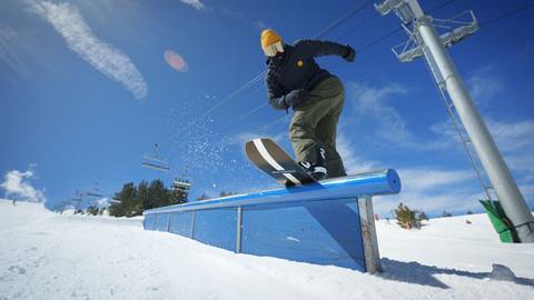 Snowboarder in black hoodie, green pants, and yellow beanie with goggles front board sliding a flat tube rail