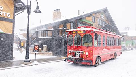 A red transportation shuttle parked outside of the main lodge of Snow Summit during a snowy day with snow falling.
