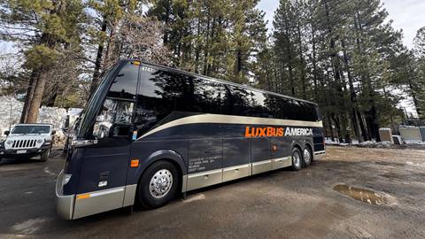 A large dark colored shuttle bus, with the company name "LuxBus America" on the side in orange and white font, in a parking lot with tall green pine trees on a sunny day with clouds in the sky.