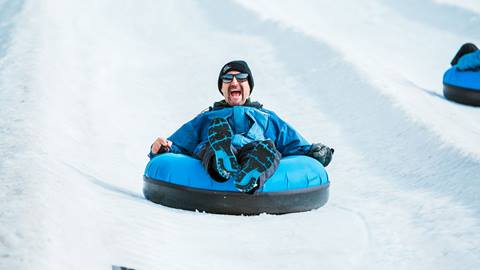 Adult in a blue jacket and black beanie with black glasses, riding a blue snow tube down a tubing lane during winter at Snow Valley's Coyote Creek Tube Park.
