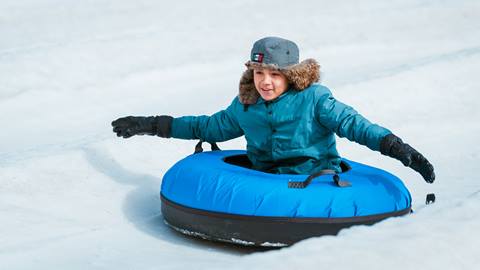 A kiddo wearing a dark teal jacket, black snow gloves, and a grey beanie with faux fur riding down the tubing lane in a blue inflatable tube at Snow Valley's Coyote Creek Tube Park.