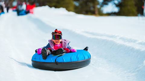 Little kiddo with a big smile, wearing pink snow gloves and goggles with a red bow in their hair, gliding down a Coyote Creek Tube Park tubing lane at Snow Valley during a sunny winter day.