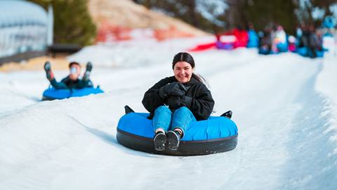 A smiling adult wearing blue pants, black long sleeve jacket, and black shoes sitting in a blue tube while gliding down a snow tubing lane at Coyote Creek Tube Park at Snow Valley.