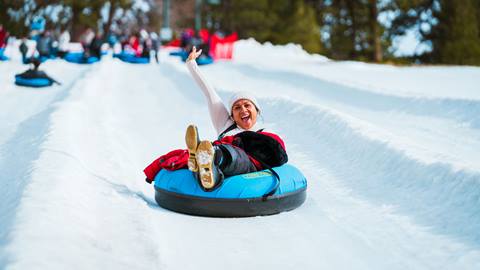 Adult in white beanie sitting in a blue winter tube and extending their right arm in the air with excitement, sliding down the lane, while guests are waiting their turn up at the top of the hill at Snow Valley's Coyote Creek Tube Park.