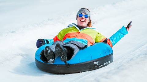 A smiling adult wearing reflective sunglasses and a bright green, orange, and blue horizonal stripped jacket while sliding down a tube at Snow Valley's Coyote Creek Tube Park during winter.