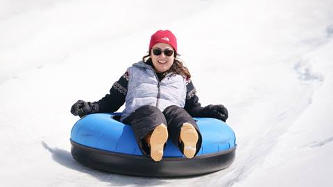Adult in a grey snow vest with black pants riding in a snow tube while zooming down a lane at Snow Valley's Coyote Creek Tube Park