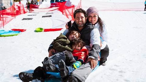 A family of four, two adults and two children, siling for the camera while sitting on a snow sled in the Snow Valley Snow Play area on a sunny winter day.