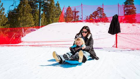 Adult with long black hair and their child with blonde hair on a snow sled at Snow Valley playing in the red fenced in Snow Play area on a sunny blue sky winter day.