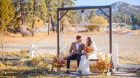 A newlywed couple, dressed in a suit and wedding gown, sitting on a detachable chairlift at Bear Mountain Golf Course on a sunny evening day with Bear Mountain in the background.