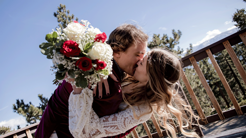Bridge and groom kissing, bride holding flowers, on deck at top of mountain