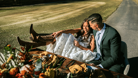 Bride sitting closely in groom's lap outdoors near a reception table