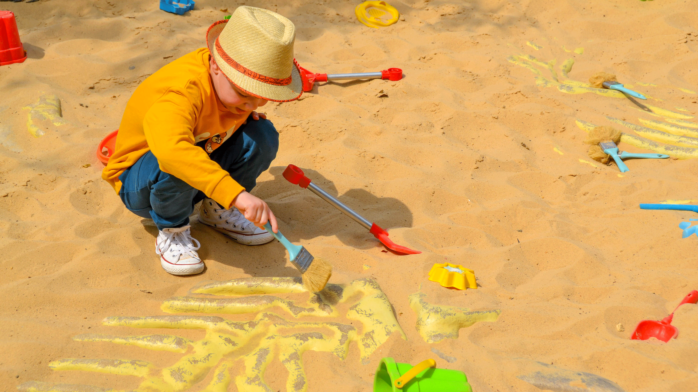 Young toddle wearing a yellow hate and a mustard color sweater with jeans holding a blue brush, uncovering fake fossils in a sandbox