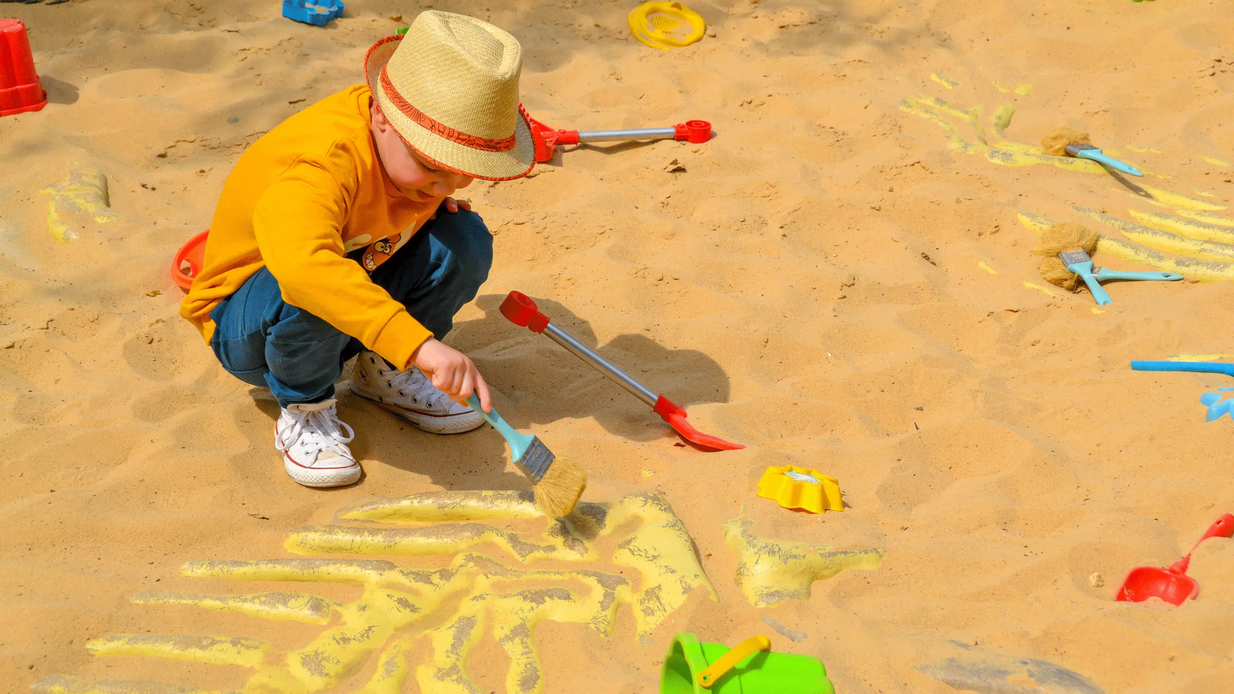 Young toddle wearing a yellow hate and a mustard color sweater with jeans holding a blue brush, uncovering fake fossils in a sandbox